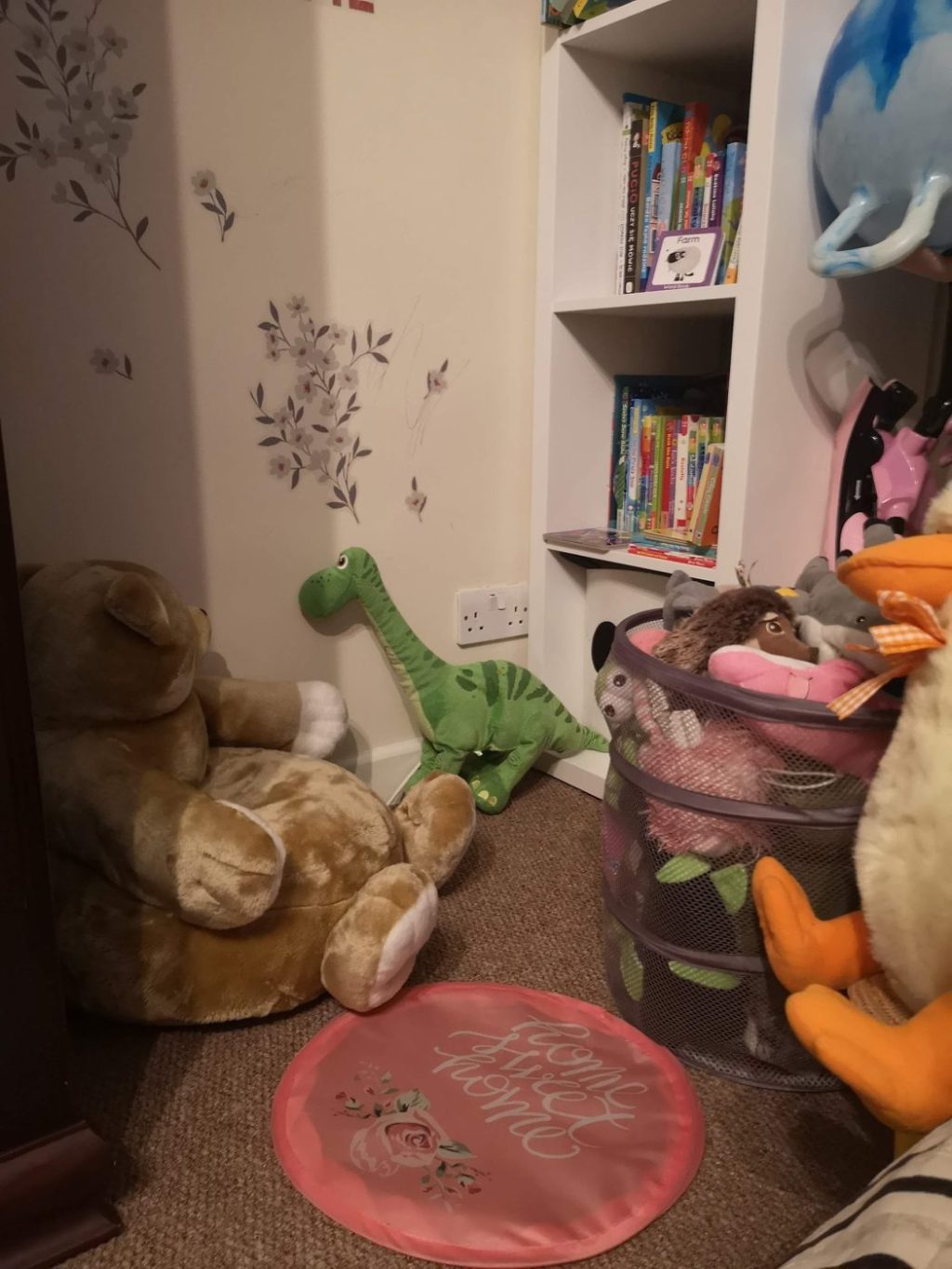 A cozy kids' reading corner with a teddy bear seat, a green dinosaur plush and a white bookshelf full of picture books